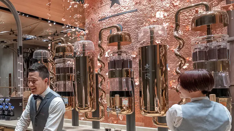 Two baristas in front of a row of cold brew silos which include intricate, curved pipes