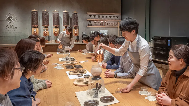 Three people serving tea to a dozen people at a long table in the AMU Inspiration Lounge
