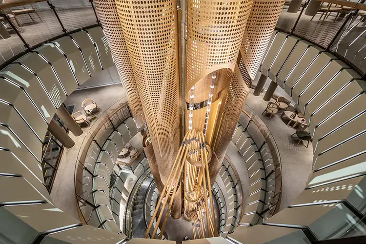 A foreshortened view looking down at the Chicago Roastery metal cask from the fourth floor to the first floor, with both clear and metal tubes transporting coffee, and a spiral escalator and some seating visible on the different floors