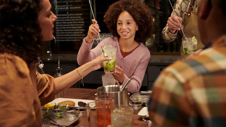 An adult woman holds a fruity beverage while a kid uses a long handled spoon to stir.