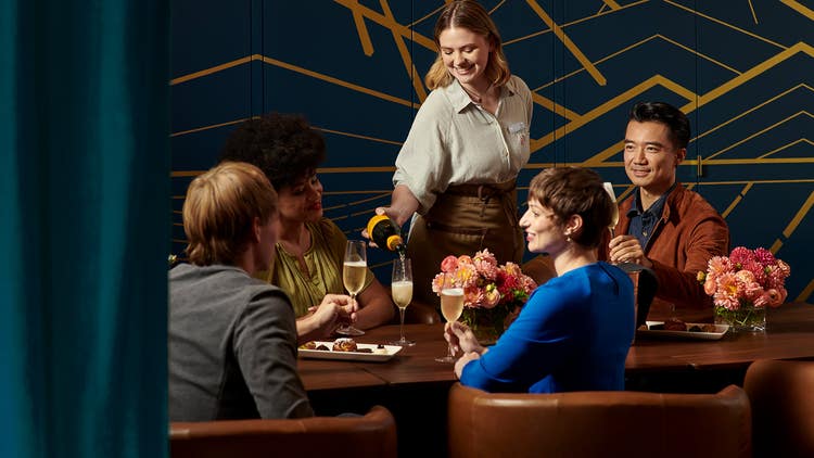 A group of people sitting around a table, enjoying champagne being poured by a bartender.