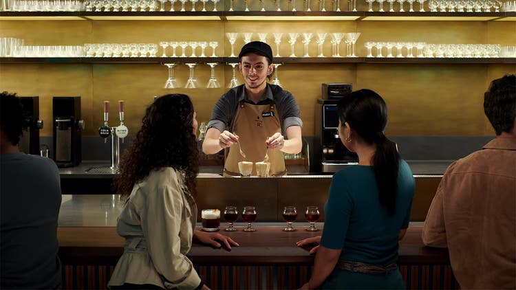 A bartender preparing beverages while two customers watch from a counter.